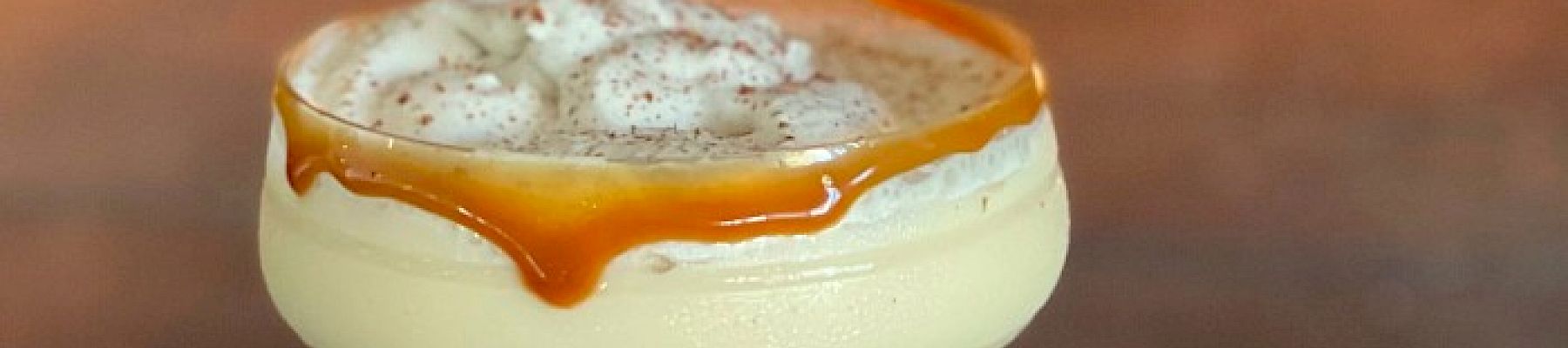A creamy cocktail in a glass with caramel drizzling down the side, topped with whipped cream and sprinkled spices, set against a background of flames.