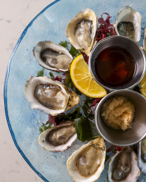 A plate of oysters arranged in a circular pattern with a lemon wedge, two condiment bowls, and garnishes on a blue plate.