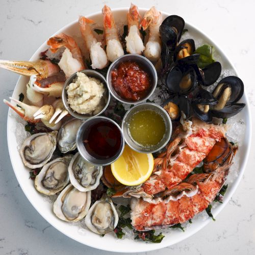 A seafood platter with shrimp, oysters, lobster, and mussels, accompanied by lemon wedges and various dipping sauces in small bowls on a white plate.