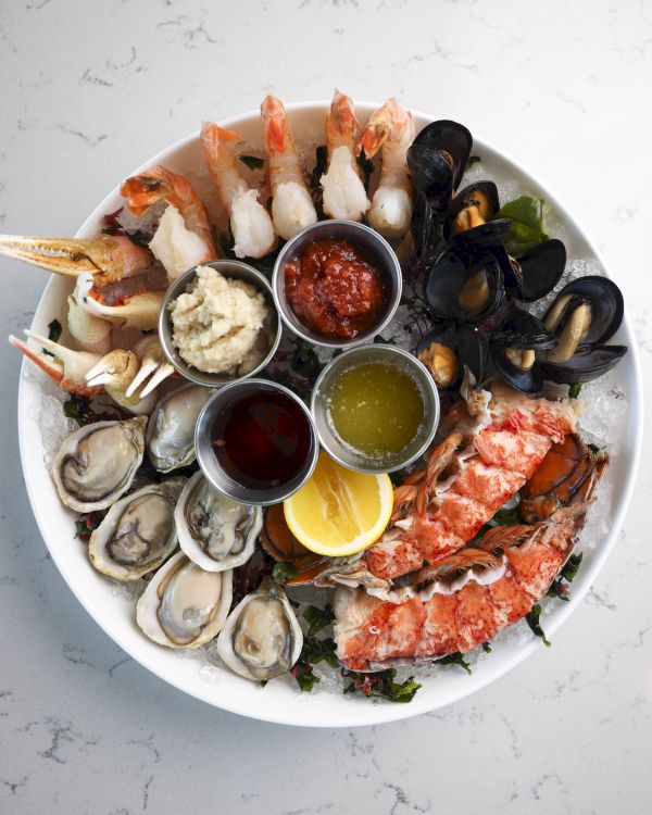 A seafood platter with shrimp, oysters, lobster, and mussels, accompanied by lemon wedges and various dipping sauces in small bowls on a white plate.