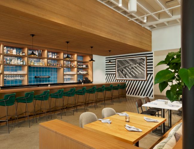 A restaurant bar with green stools, blue tile backsplash, and industrial-style pendant lights. Dining tables with white napkins and plants are visible.