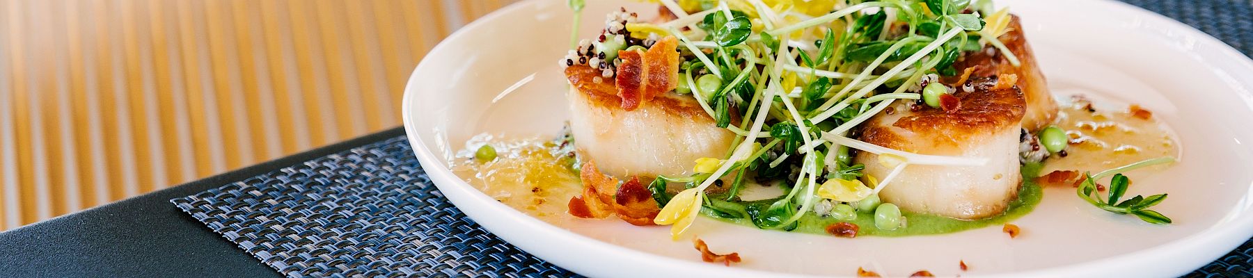 The image shows a plate of gourmet food, possibly seared scallops, garnished with greens and microgreens, placed on a blue woven placemat on a table.