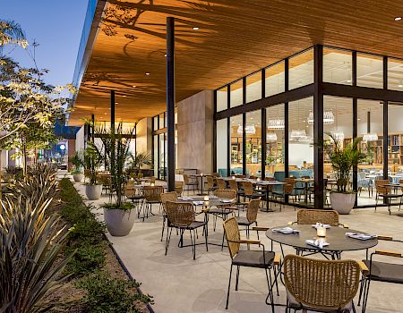 An outdoor restaurant seating area with tables, chairs, and plants in pots. Adjacent is an illuminated indoor dining space with large glass windows.