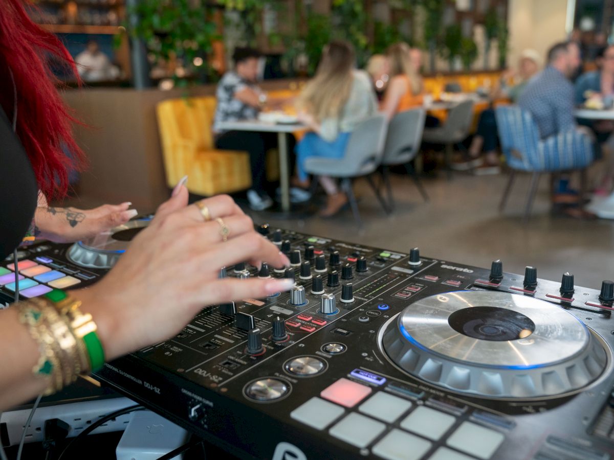A DJ with red hair is mixing music on a console in a lively restaurant with people dining in the background.