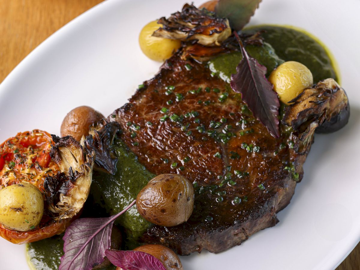 A delicious-looking plated dish featuring a hearty steak or roast, garnished with roasted potatoes and greens on a white oval plate.