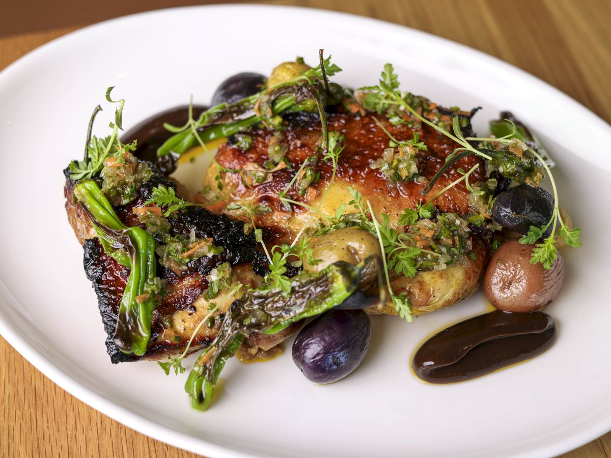 A plated dish featuring grilled chicken with herbs, potatoes, green vegetables, and a dark sauce garnish on a white plate.